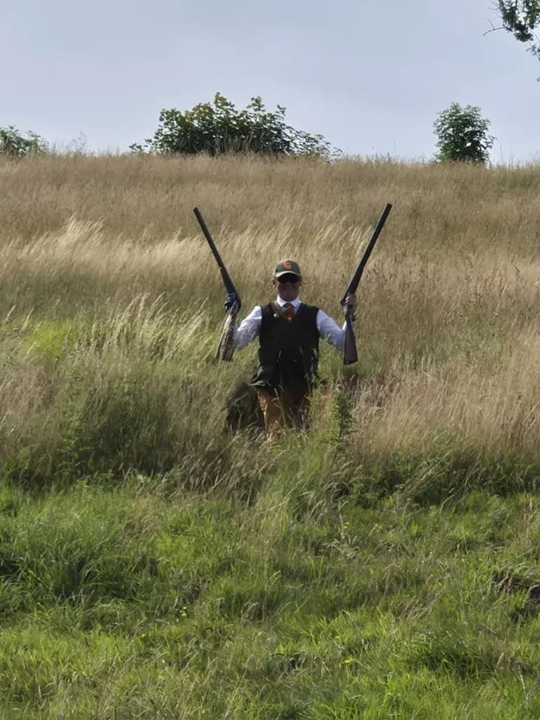 Chasseur brandissant fièrement deux fusils dans les herbes hautes après une battue simulée au Domaine de La Planquette